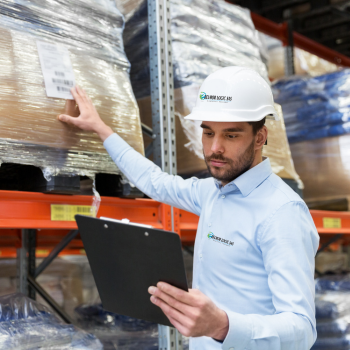 primer plano de un hombre joven con casco de protección, revisando la etiqueta de una caja en una bodega de almacenamiento, con una tabla para inventarios en su mano izquierda.